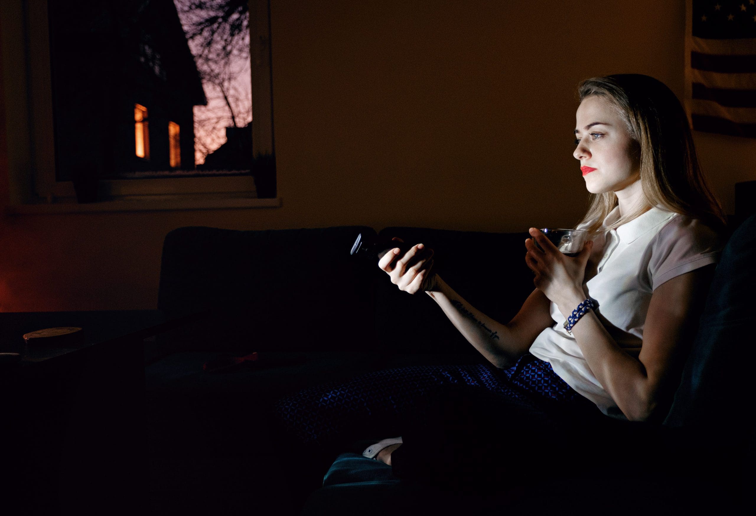 Side view of young female sitting on sofa in twilight with mug and switching channels of tv. *NOTE- THESE IMAGES WERE SHOT WITH A HIGH ISO AND EXHIBIT HIGH NOISE.